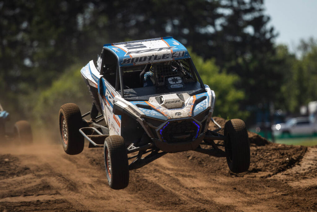 A Maxxis off-road racing UTV jumps through the air on a dirt track.