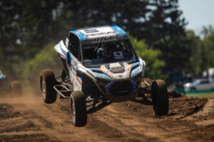 A Maxxis off-road racing UTV jumps through the air on a dirt track.