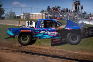 A blue Maxxis off-road racing truck jumps through the air on a dirt track.