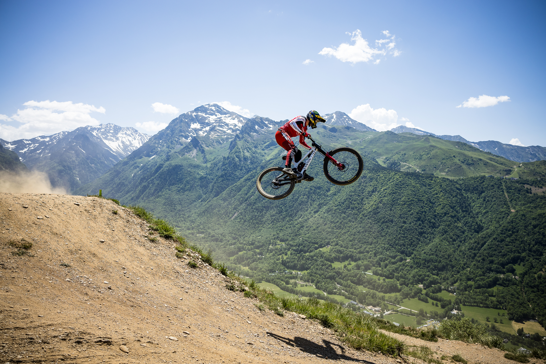 Downhill rider airborne over a jump with alpine mountains in the background, showcasing Maxxis Minion DHX tire performance.