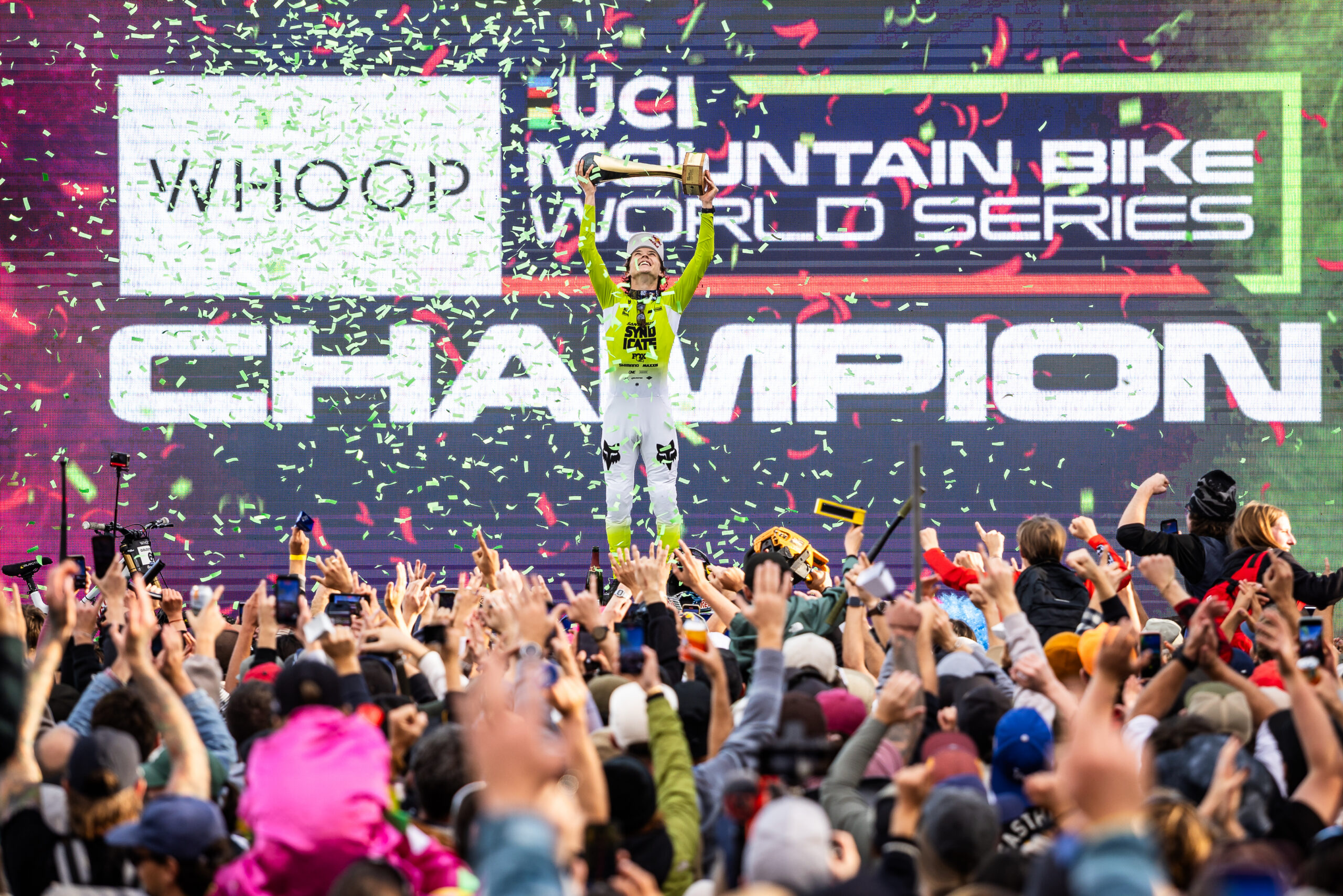 Downhill mountain bike champion lifts a trophy on the World Cup podium as fans celebrate below.