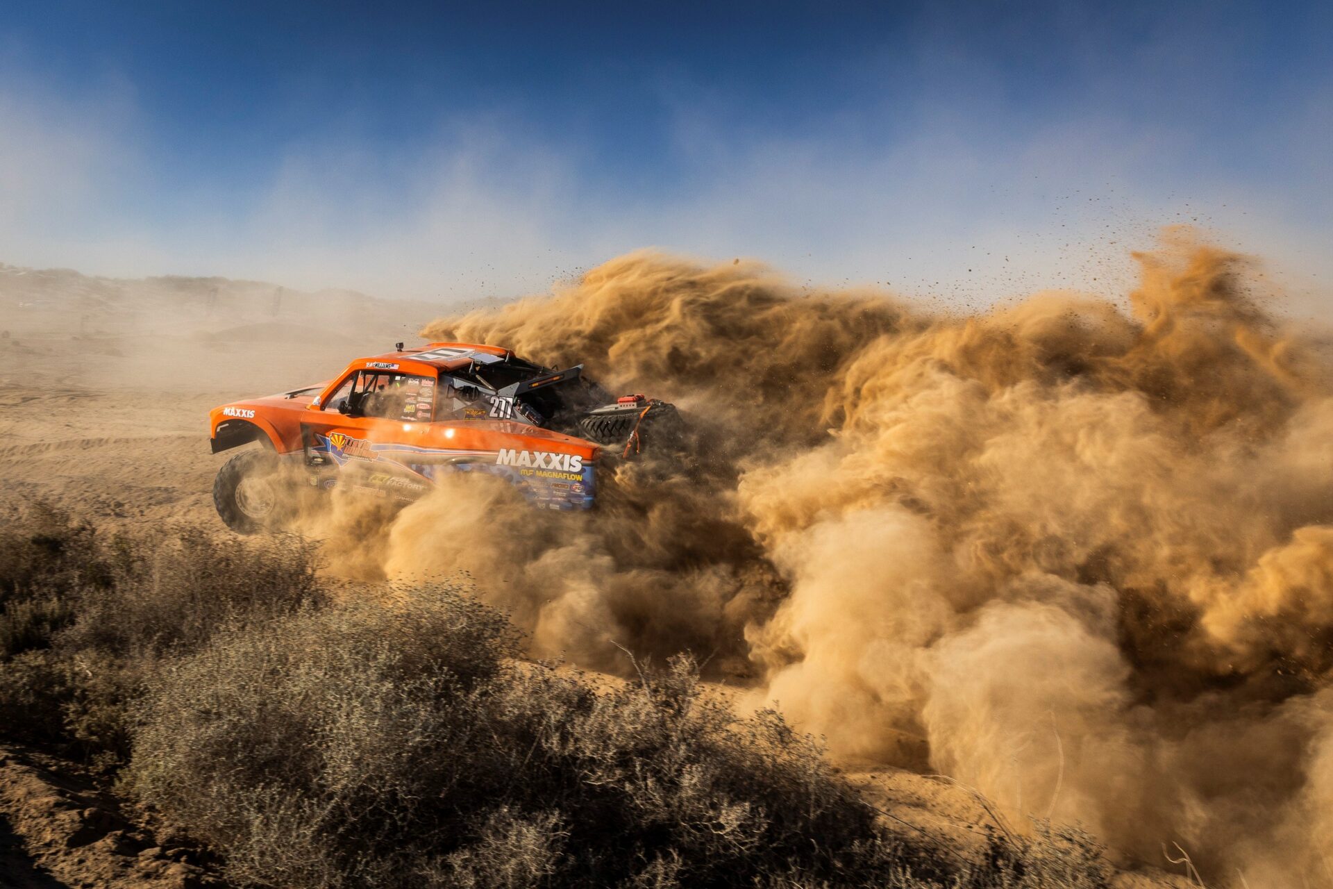 A bright orange off-road racing truck speeds through the desert, creating a massive, swirling cloud of golden dust that fills the frame.